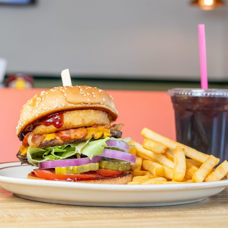 A burger with onion rings, bacon, cheese, lettuce, onion, pickles, and tomato, with fries and a drink.