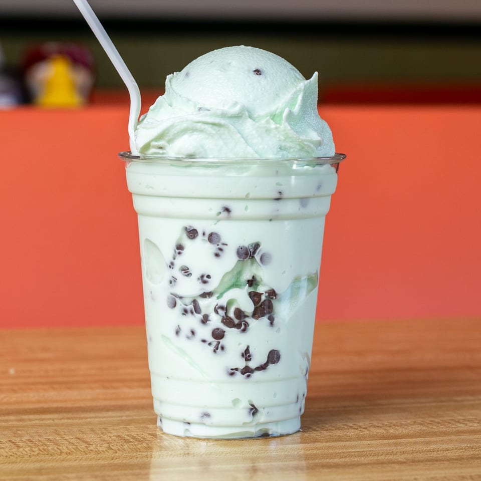 Mint chocolate chip ice cream in a clear plastic cup with a spoon on a wooden table.