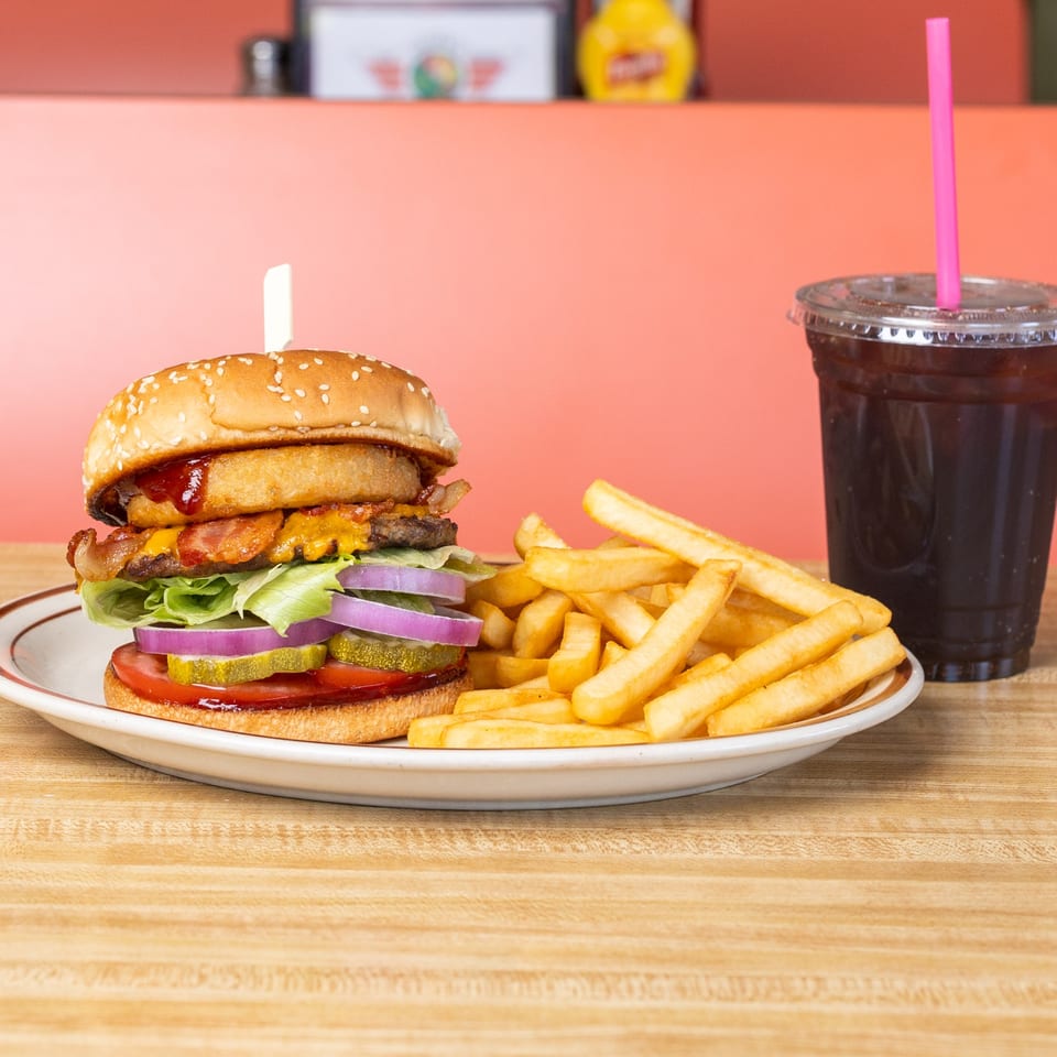 A bacon cheeseburger with onion rings, fries, and a soda with a pink straw on a diner table.