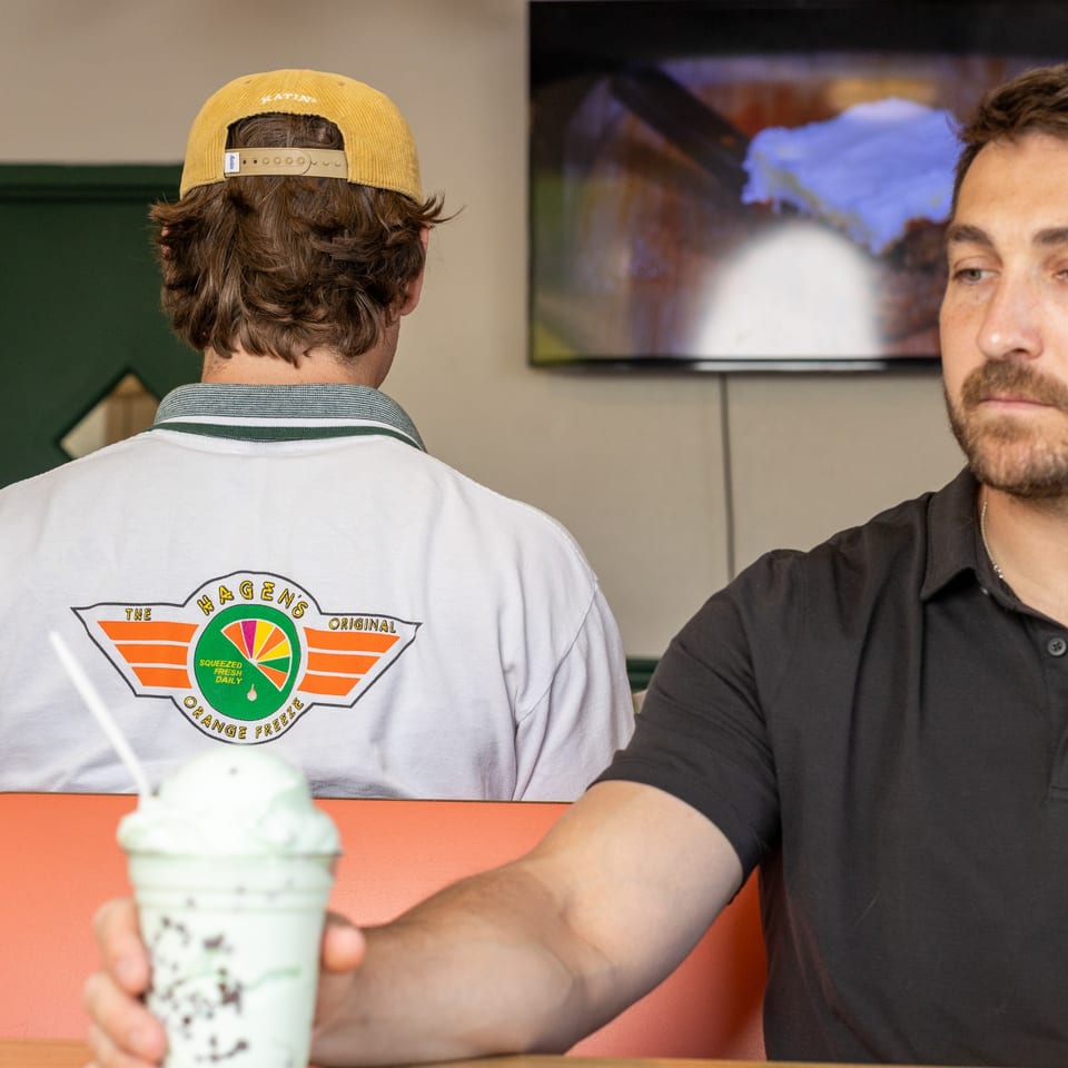 Two men sit at a booth, one holding a mint chocolate chip milkshake, watching TV.